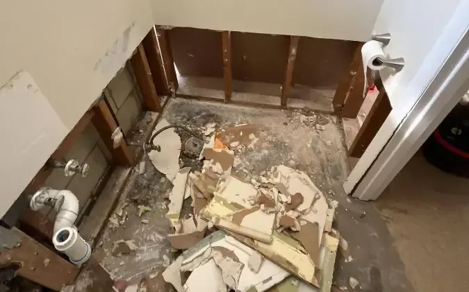 Removed wallboard lies on a bathroom floor below exposed studs and plumbing connections.