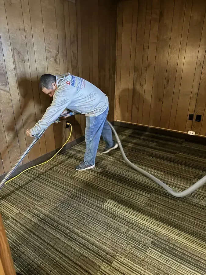 A technician running an extraction hose and wand across a wet carpet in a wood-paneled room