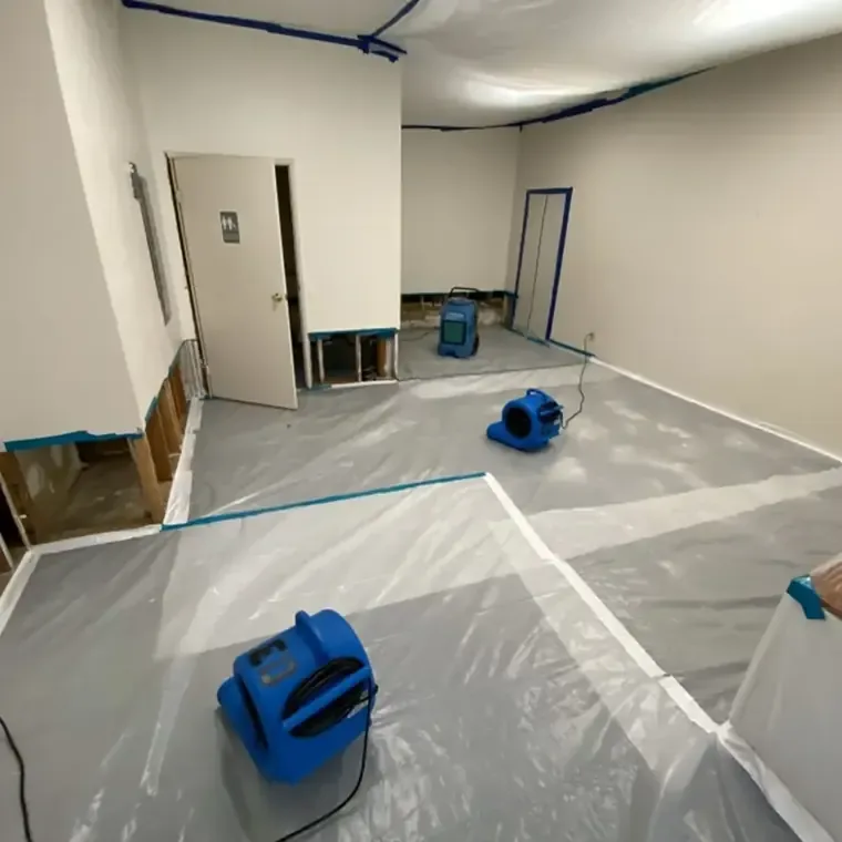 Blue air movers stand on plastic covered flooring beside walls with their lower sections opened.
