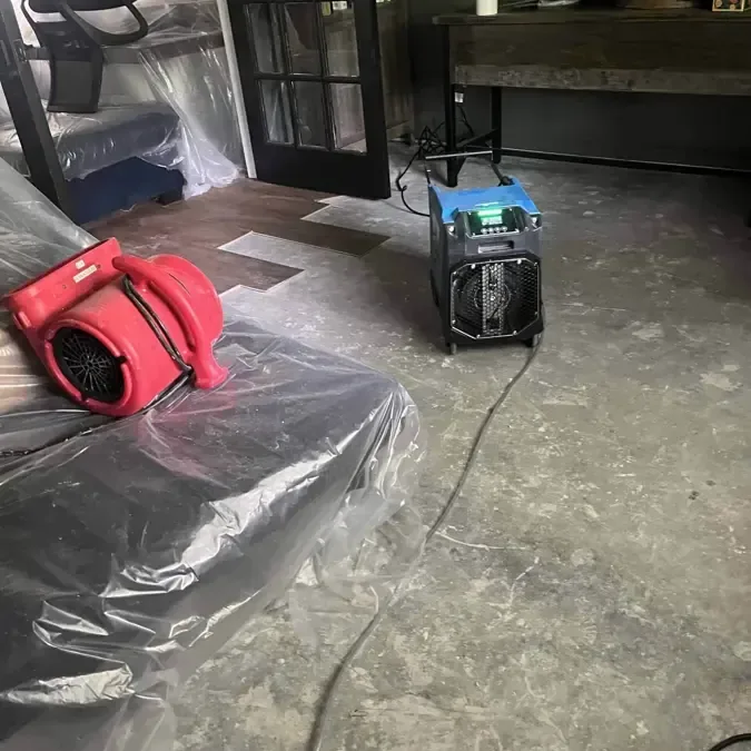 A red air mover and a dehumidifier stand beside plastic sheeting on an exposed floor.