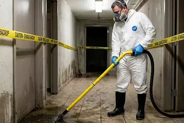 Worker in protective suit and respirator extracting a sewage backup behind caution tape